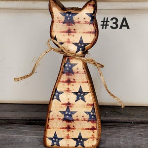 May include: Wooden cat figurine with a patriotic design. The cat is painted with a distressed American flag pattern, featuring blue stars and red stripes. A twine bow is tied around the neck. The text "#3A" is visible.
