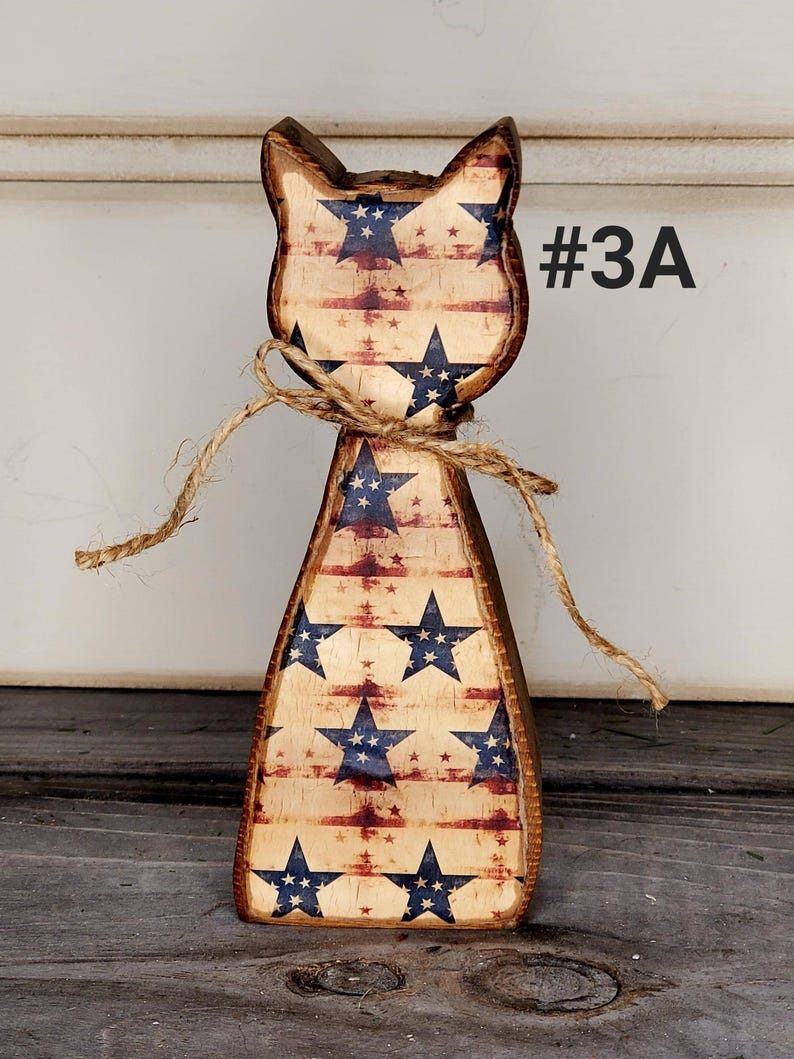 May include: Wooden cat figurine with a patriotic design. The cat is painted with a distressed American flag pattern, featuring blue stars and red stripes. A twine bow is tied around the neck. The text "#3A" is visible.