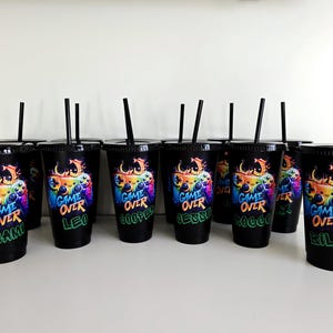 May include: A collection of black plastic tumblers with lids and black straws. Each cup features a colourful graphic of a video game controller with the text "GAME OVER" and a personalised name printed in a vibrant, graffiti-style font.