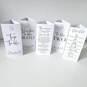 May include: Four white triangular table cards with black text. The cards are labelled "Top Table", "Vendor Table Buffet", "Table Two", and "Let's Eat". The "Let's Eat" card lists the names of different desserts.