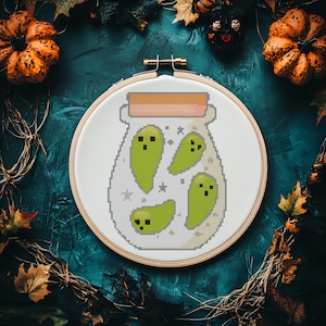 Peut inclure: Un cercle de broderie au point de croix avec un motif pixélisé de fantômes verts dans un bocal. La broderie est posée sur un fond bleu canard foncé avec des citrouilles, des feuilles et une araignée.