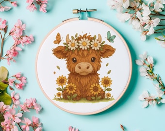 Op de afbeelding: Een ronde borduurring met een cartoon illustratie van een Schotse Hooglandkoe. De bruine koe draagt een madeliefjeskrans en is omringd door zonnebloemen en vlinders. Op een lichtblauwe achtergrond, met roze en witte bloemen.