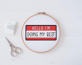 May include: A white embroidery hoop with a red name tag that says "Hello I'm Doing My Best" in black text. There is a spool of white thread and a pair of silver embroidery scissors on a white surface.