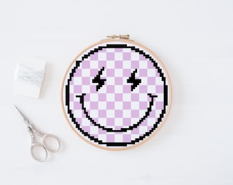 May include: A pink and white checkered smiley face with lightning bolt eyes, embroidered on a round wooden hoop.