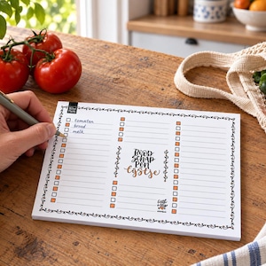 May include: A rectangular white notepad with a black decorative border and orange checkboxes. The center features the text 'Boodschappenlijstje' in black and orange script. The notepad has lined sections for writing, with 'tomaten', 'brood', and 'melk' written in the first column.