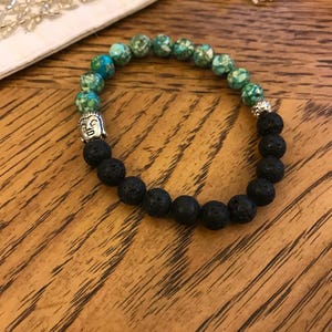 Puede incluir: Un brazalete de piedra de lava negra y piedra preciosa verde con un dije de cabeza de Buda plateado.