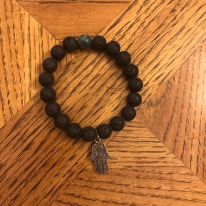 May include: A black lava rock bracelet with a green stone bead and a silver hamsa charm.