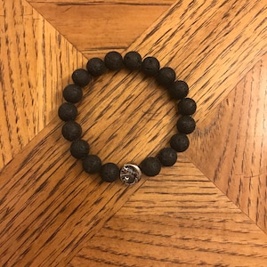Może przedstawiać: Bransoletka z czarnych koralików z lawy wulkanicznej z srebrnym zawieszką. Bransoletka leży na drewnianej powierzchni.