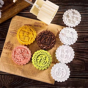 Puede incluir: Una colección de coloridos pasteles de luna redondos con diseños intrincados y caracteres chinos. Los pasteles de luna son en tonos de amarillo, marrón, rosa y verde, y están dispuestos sobre un papel vintage con texto. Un molde blanco está en el fondo.