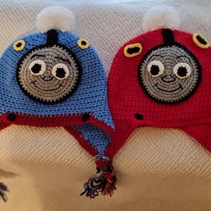 May include: Two crocheted hats, one blue and one red, each with a smiling train face. The hats have ear flaps, tassels, and a white pom-pom. The train faces are grey with black and white details.