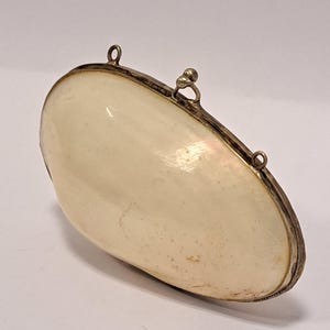 May include: A clam-shaped clutch bag with a pearlescent, off-white surface and a silver-coloured metal frame. The bag has a small clasp and two small loops attached to the frame.