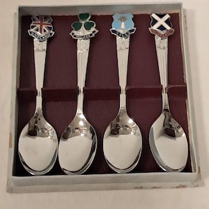 Könnte beinhalten: Ein Set aus vier silberfarbenen Souvenir-Löffeln in einer Schachtel. Jeder Löffel hat oben ein dekoratives Emaille-Emblem, das England, Irland, Wales und Schottland darstellt, wobei die jeweiligen Namen auf der Basis eingraviert sind.