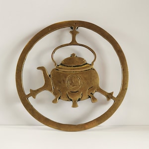 Puede incluir: Un calienta-tetera de latón con un diseño circular. El calienta-tetera presenta un recorte detallado de una tetera con tapa y asa.