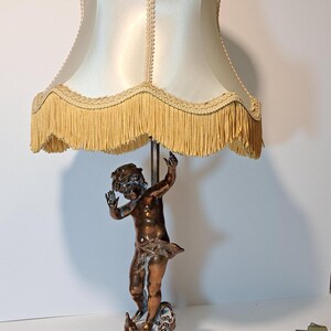 May include: A table lamp featuring a bronze-toned cherub figure as the base, standing on a circular pedestal. The lamp is topped with a cream-colored satin fabric shade, which is finished with a decorative braided trim and a long fringe border.