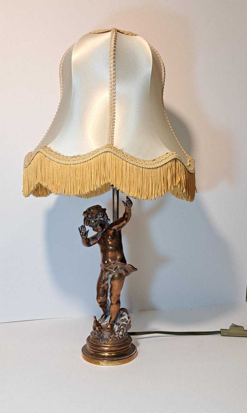 May include: A table lamp featuring a bronze-toned cherub figure as the base, standing on a circular pedestal. The lamp is topped with a cream-colored satin fabric shade, which is finished with a decorative braided trim and a long fringe border.