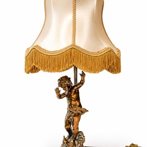 May include: A table lamp featuring a bronze-toned cherub figure as the base, standing on a circular pedestal. The lamp is topped with a cream-colored, bell-shaped fabric shade finished with a decorative gold-colored fringe trim along the bottom edge.