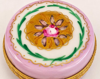 以下が含まれることがあります： 淡い紫色の縁取りと、手描きのピンクと茶色の花柄が描かれた中央の金色のメダルが特徴の丸い磁器製小物入れ。蓋には緑の葉のモチーフと金色の縁取りが施されており、ベースには金色の金属製ヒンジが付いています。