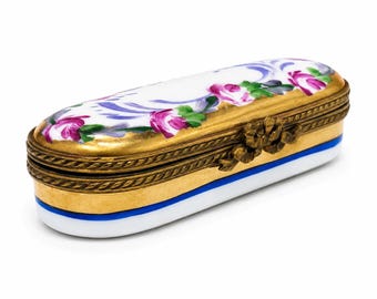 May include: An oval porcelain trinket box featuring a white lid with hand-painted pink roses and purple scrollwork. The box has a gold-toned metal rim with a decorative bow clasp and a white base accented by a single horizontal blue stripe.