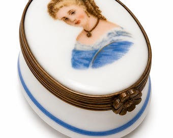 May include: An oval porcelain trinket box with a brass hinge and a flower-shaped clasp. The lid features a hand-painted portrait of a person with curly hair, wearing a blue dress and a dark necklace. A thin blue line encircles the white base.