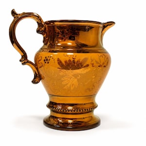 May include: A metallic copper-colored glass pitcher featuring a reflective finish and an ornate, curved handle. The body of the pitcher is decorated with an etched floral pattern and a beaded band detail around the base, with a small pouring spout.