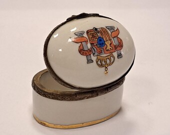 May include: A white porcelain trinket box with a gold trim and a hinged lid. The lid features a colorful painted design of a coat of arms with a crown and the words "Plus Ultra".