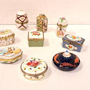 May include: A collection of eight small porcelain trinket boxes featuring various shapes and designs, including floral patterns, a nautical scene, a dark blue box with orange koi fish, and gold-toned metal hinges and clasps on each piece.