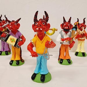 May include: A set of five red devil figurines, each with black horns, standing on green circular bases. The figures are dressed in colourful shirts and trousers, and each holds a different musical instrument, including an accordion, a triangle, and horns.