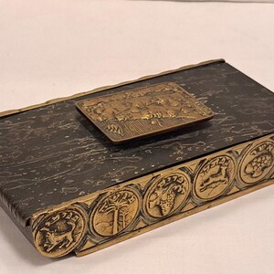 May include: A rectangular, antique box with a dark, textured surface and bronze-colored trim. The lid features a bronze plaque with a detailed relief scene. The box's edge is adorned with circular relief designs.