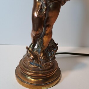 May include: A metallic bronze-toned table lamp base featuring a sculpted figure of a cherub standing on a circular pedestal. The figure is draped in fabric around the waist and stands beside a small bird, with a black electrical cord extending outward.