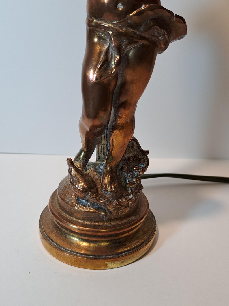 May include: A metallic bronze-toned table lamp base featuring a sculpted figure of a cherub standing on a circular pedestal. The figure is draped in fabric around the waist and stands beside a small bird, with a black electrical cord extending outward.