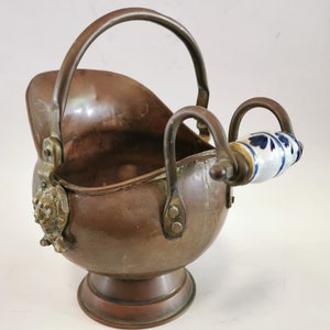 May include: A copper coal scuttle with a decorative handle and a porcelain knob. The scuttle has a round base and a curved body.