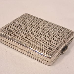 Könnte beinhalten: Ein rechteckiges Silberetui mit Scharnierdeckel. Das Etui ist mit einem sich wiederholenden Muster aus horizontalen Linien und ovalen Formen verziert. Der Verschluss befindet sich auf der rechten Seite. Das Etui wird wahrscheinlich zum Tragen kleiner Gegenstände verwendet.