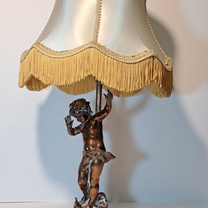 May include: A table lamp featuring a bronze-toned figure of a cherub standing on a circular base. The lamp has a cream-colored satin shade with decorative braided trim and a long, hanging fringe along the bottom edge of the lampshade.
