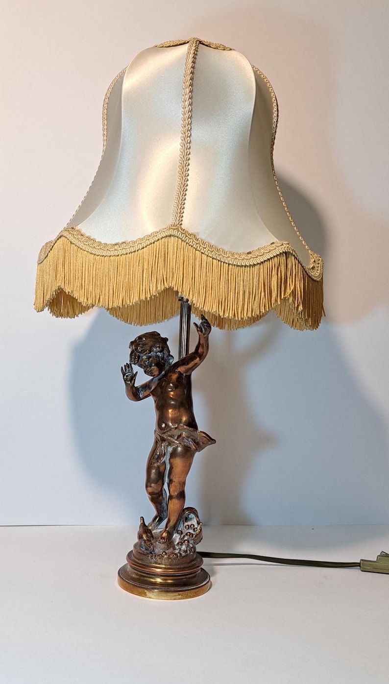 May include: A table lamp featuring a bronze-toned figure of a cherub standing on a circular base. The lamp has a cream-colored satin shade with decorative braided trim and a long, hanging fringe along the bottom edge of the lampshade.