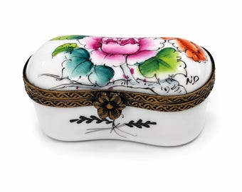 May include: An oval white porcelain trinket box with a hinged lid featuring a hand-painted pink peony and green leaves. The box has a decorative bronze-toned metal rim with a flower-shaped clasp and a small black floral motif on the front side.