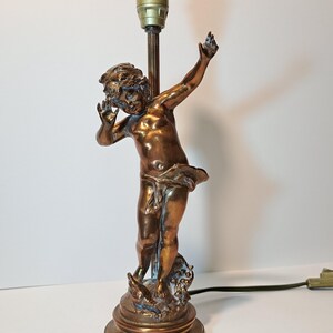 May include: A bronze-toned metal table lamp base featuring a sculpted figure of a child with arms raised. The figure stands on a circular base with decorative elements at the feet. A vertical rod extends upward from the figure to a light bulb socket.