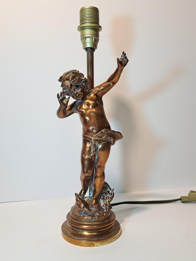 May include: A bronze-toned metal table lamp base featuring a sculpted figure of a child with arms raised. The figure stands on a circular base with decorative elements at the feet. A vertical rod extends upward from the figure to a light bulb socket.