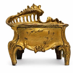 Puede incluir: Una pequeña caja decorativa pintada en dorado con un respaldo curvo y ornamentado con listones verticales y cuatro patas talladas. El panel frontal muestra un sutil relieve floral, con zonas visibles de acabado desgastado.