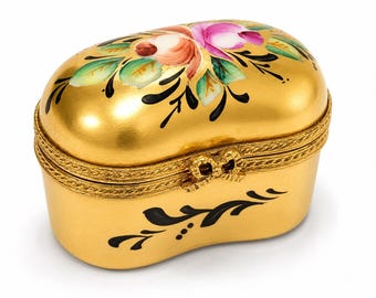 May include: An oval gold-colored trinket box with a hinged lid. The lid features a hand-painted floral design in pink, orange, and green. The base and lid are decorated with black painted flourishes and a textured, rope-like metal trim.