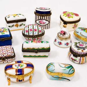 May include: A collection of twelve small porcelain trinket boxes featuring various shapes, including rectangular, oval, and heart-like forms. Each box is decorated with floral patterns, stripes, or geometric designs and includes ornate metal hinges.