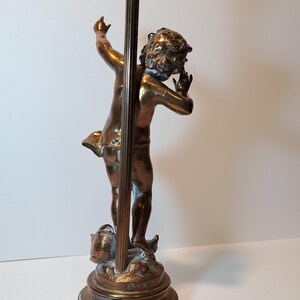 May include: A bronze-toned table lamp base featuring a sculpted figure of a child standing on a circular pedestal. The figure is positioned with one arm raised and the other near the head. A vertical metal rod extends upward from the base.