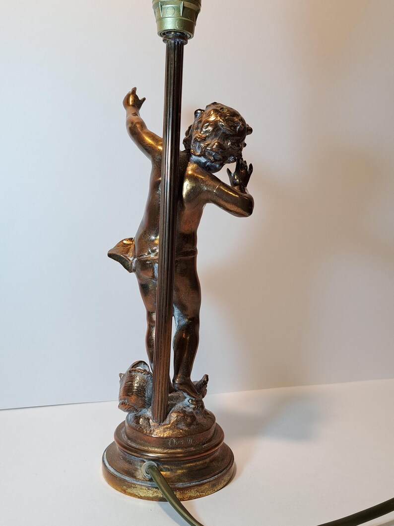 May include: A bronze-toned table lamp base featuring a sculpted figure of a child standing on a circular pedestal. The figure is positioned with one arm raised and the other near the head. A vertical metal rod extends upward from the base.