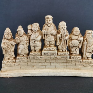 以下が含まれることがあります： 日本の神話の七福神をモチーフにした、小さな白い陶器の置物7体セット。それぞれの置物は、ポルトガル語が刻まれた小さなレンガの台座の上に立っています。言葉は、Musica、Justi&ccedil;a、Fortuna、Felicidade、Sabedoria、Boa Saude、Juventudeです。
