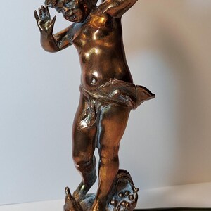 May include: A bronze-toned table lamp featuring a sculpted figure of a cherub with arms raised. The figure stands on a circular base with decorative elements at the feet. A vertical metal rod extends upward from behind the figure to support a light fixture.