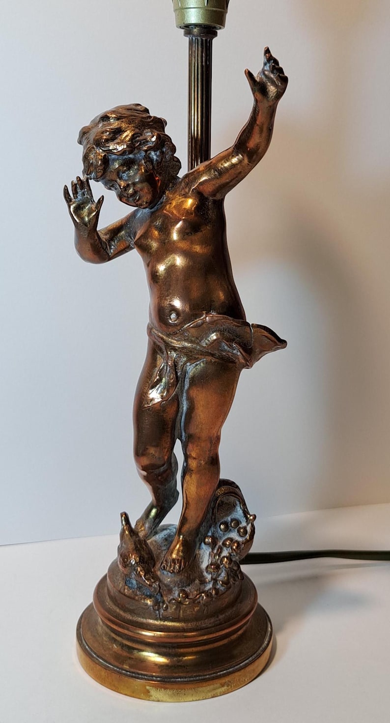May include: A bronze-toned table lamp featuring a sculpted figure of a cherub with arms raised. The figure stands on a circular base with decorative elements at the feet. A vertical metal rod extends upward from behind the figure to support a light fixture.