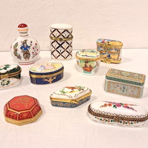 Puede incluir: Una colección de diez pequeñas cajas de porcelana para baratijas y un frasco de perfume, cada uno con intrincados diseños pintados a mano, bisagras de metal dorado y cierres. Las formas incluyen modelos ovalados, rectangulares, octogonales y cilíndricos.