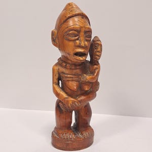 May include: A carved ivory sculpture of a standing figure holding a smaller figure. The sculpture is a traditional African art piece.