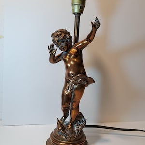 May include: A bronze-toned table lamp base featuring a sculpted figure of a cherub standing on a circular pedestal. The figure has arms raised and is draped in fabric. A vertical metal rod extends upward from behind the figure to a light socket.
