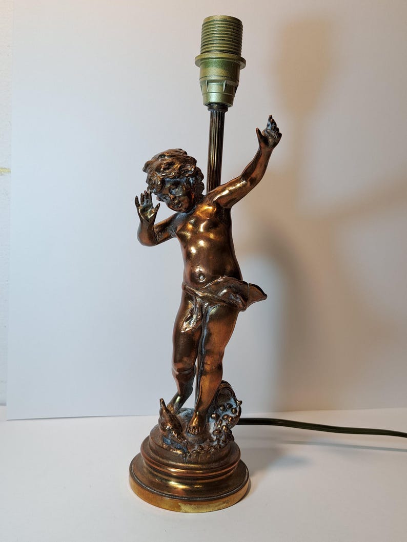 May include: A bronze-toned table lamp base featuring a sculpted figure of a cherub standing on a circular pedestal. The figure has arms raised and is draped in fabric. A vertical metal rod extends upward from behind the figure to a light socket.