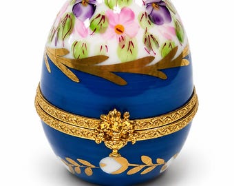 May include: A porcelain egg-shaped trinket box featuring a white top section painted with pink and purple flowers and green leaves. The bottom section is deep blue with gold leaf accents and a decorative gold metal hinge and clasp.